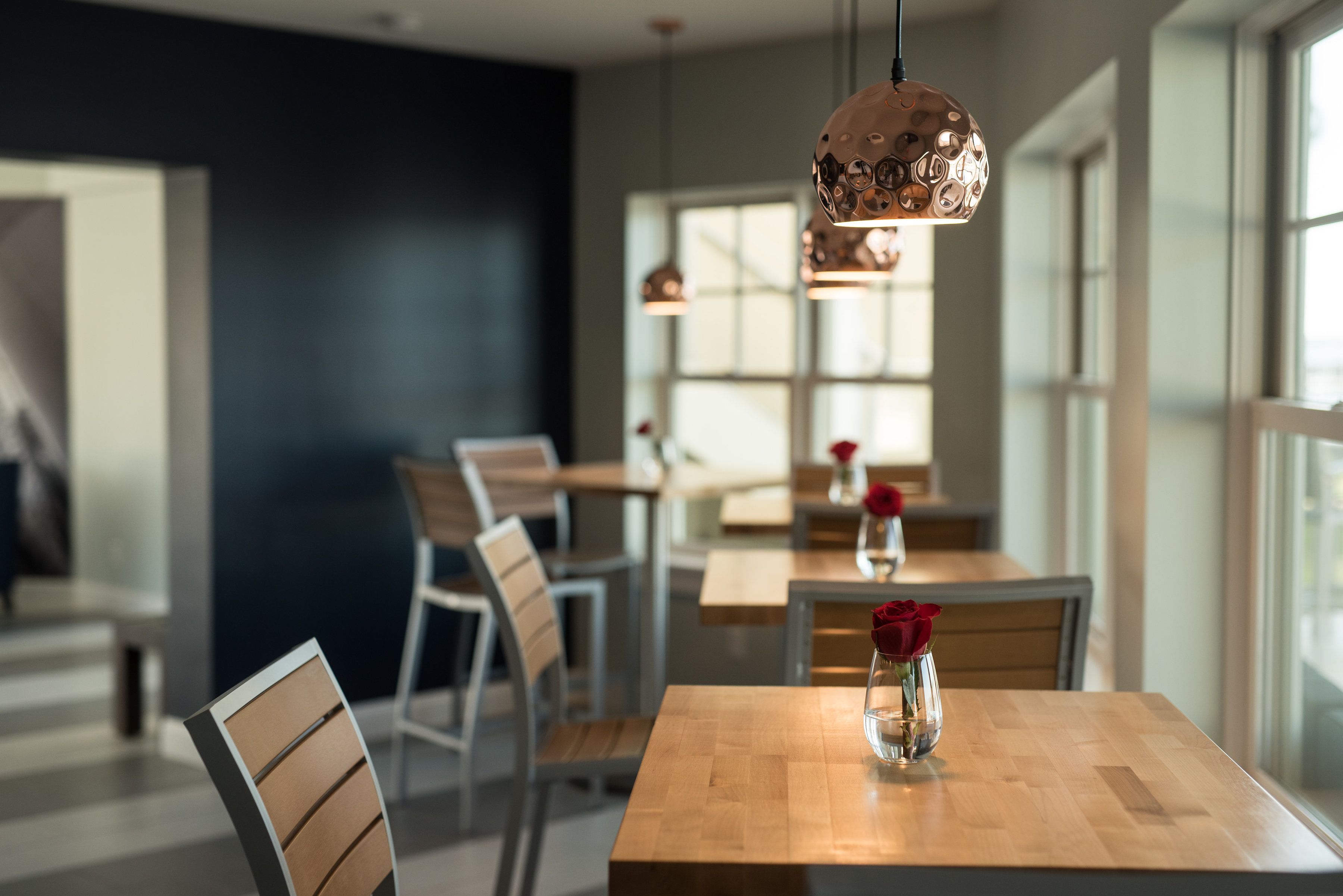 Un espace de restauration moderne avec des tables et des chaises en bois, un vase contenant une rose rouge et des luminaires suspendus.