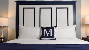 Una cama bien tendida con sábanas blancas, cabecera azul marino y un cojín con una 'M' grande.