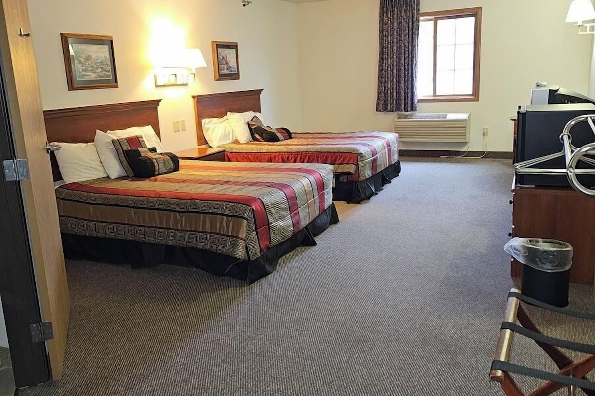 A hotel room with two beds, a desk, a chair, a window with curtains, and a ceiling-mounted air conditioning unit.