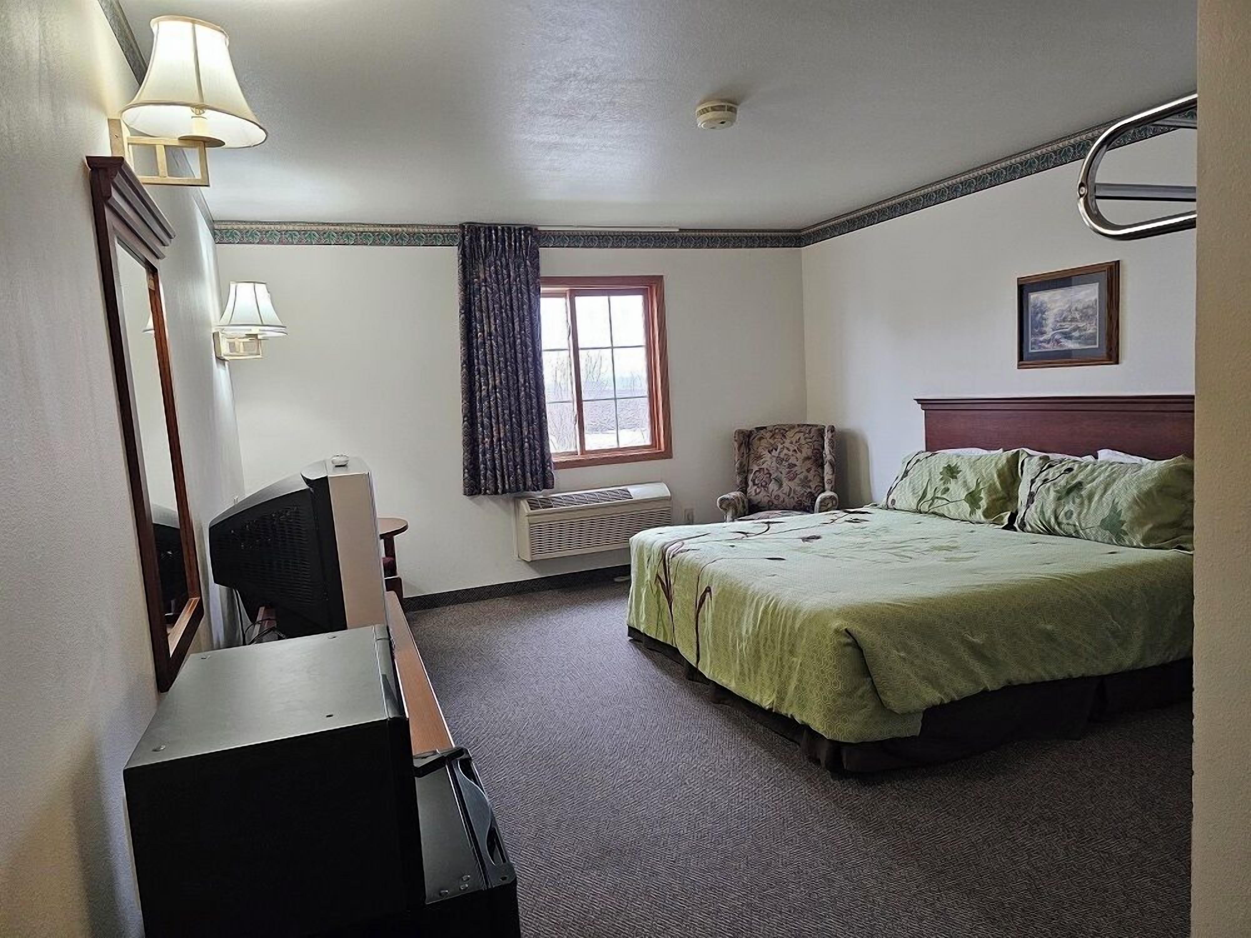 Una habitación de hotel con cama, una silla, escritorio, televisor, ventana con cortinas y un cuadro en la pared.