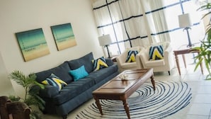 A living room with a dark blue sofa, two armchairs, a coffee table, and a patterned rug. There are two wall-mounted paintings and a window with curtains. - Encantada Resort by Home 2U (Kissimmee)
