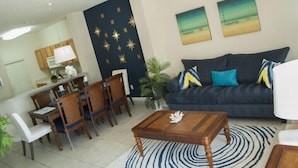 A living room with a dark blue sofa, wooden coffee table, and a dining area with wooden chairs and a table. There are two framed pictures on the wall and a ceiling fan. - Encantada Resort by Home 2U (Kissimmee)