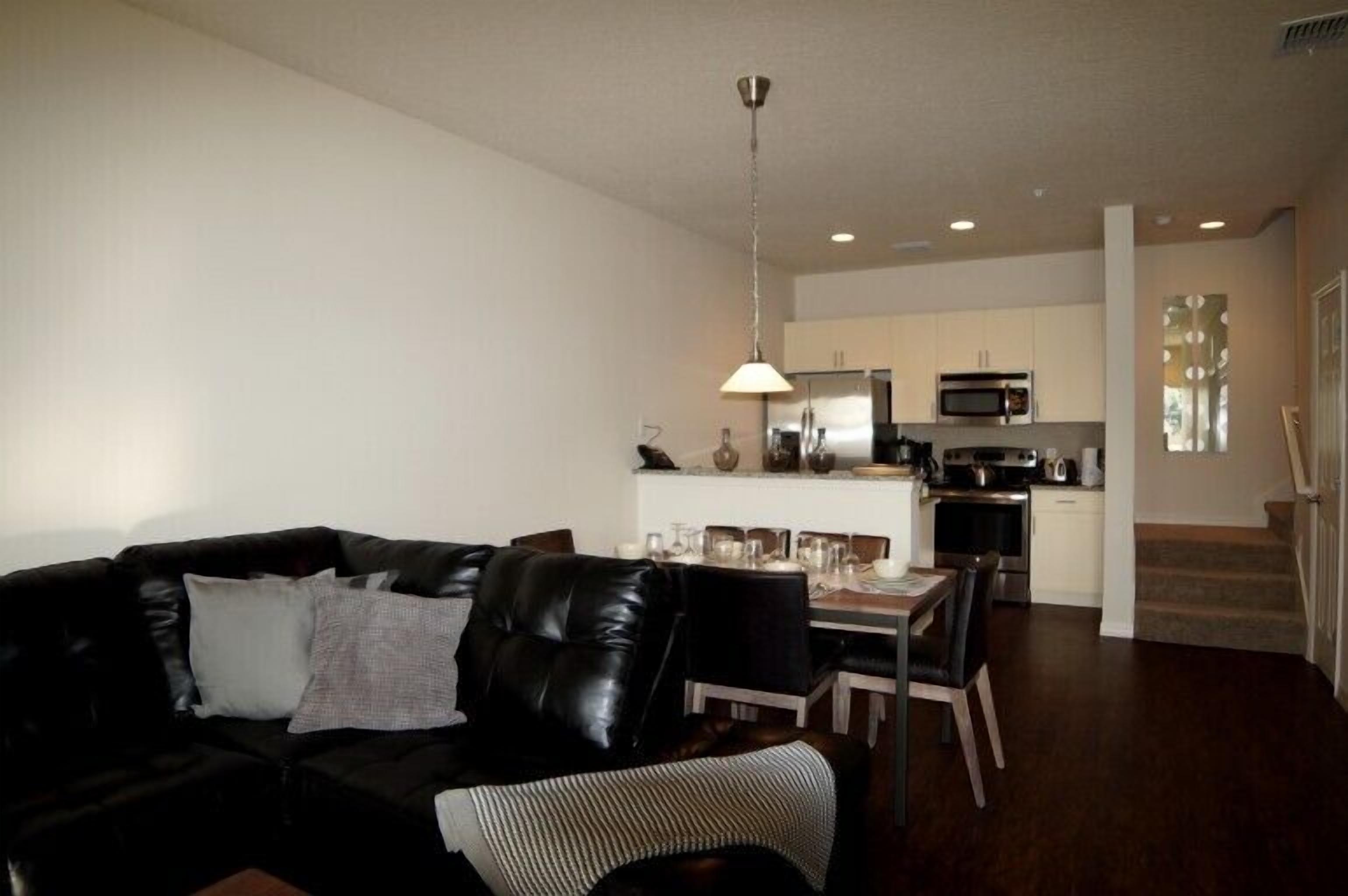 A modern living room with a black leather sofa, dining table, and chairs. A kitchen with white cabinets and appliances is visible in the background.