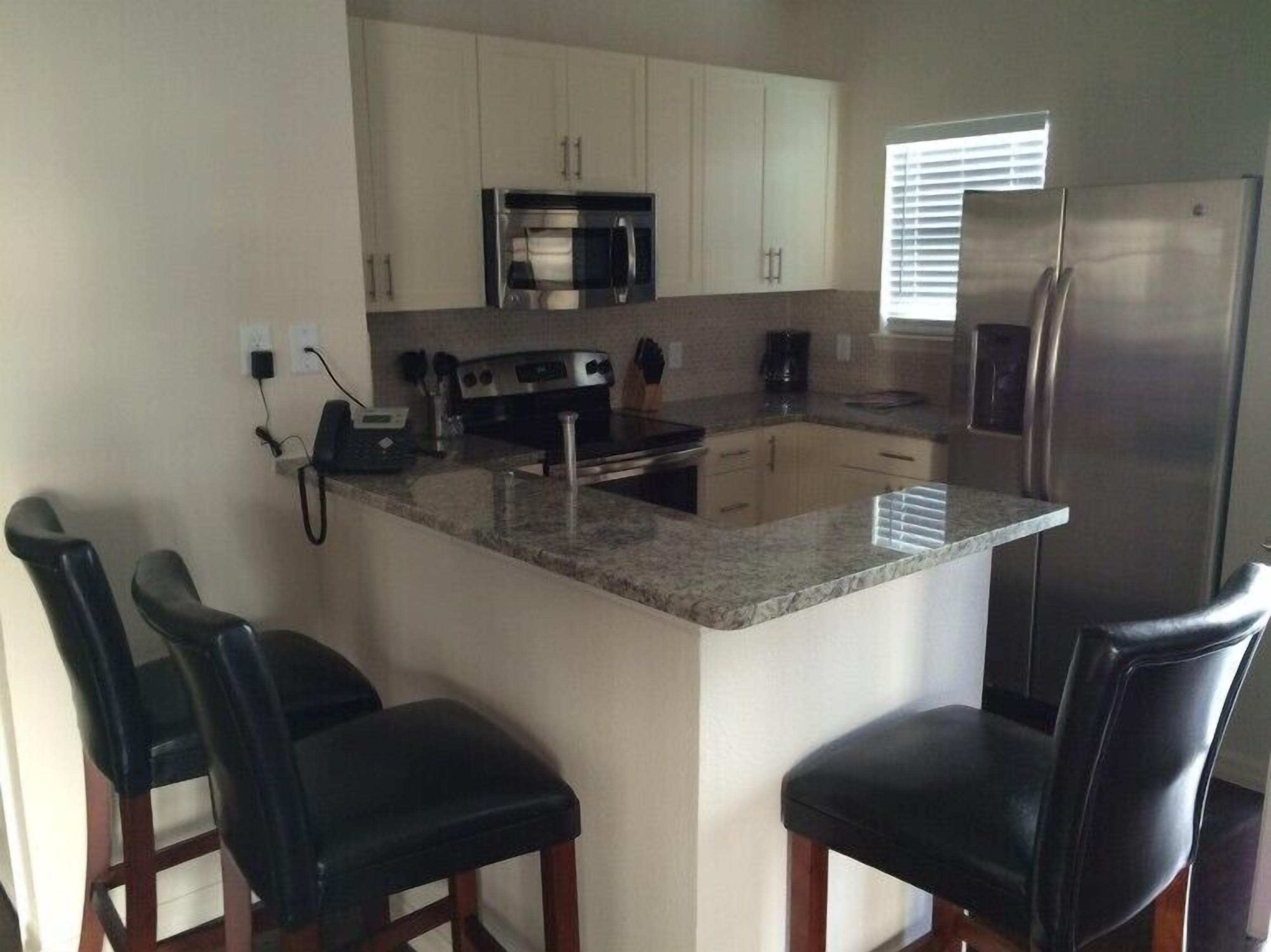 A kitchen with white cabinets, a stainless steel refrigerator, a microwave, a stove, and a coffee maker. There are three black bar stools and a phone on the counter.