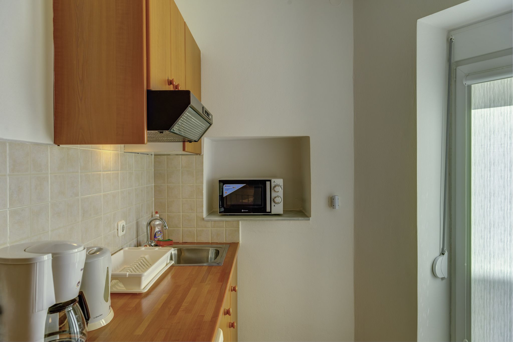 Een kleine keuken met een magnetron, spoelbak en houten keukenkastjes.