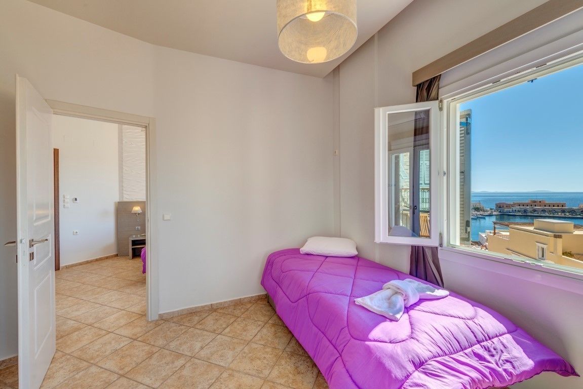 A small bedroom with a bed, a window with a view of buildings and the sea, and a door leading to another room.