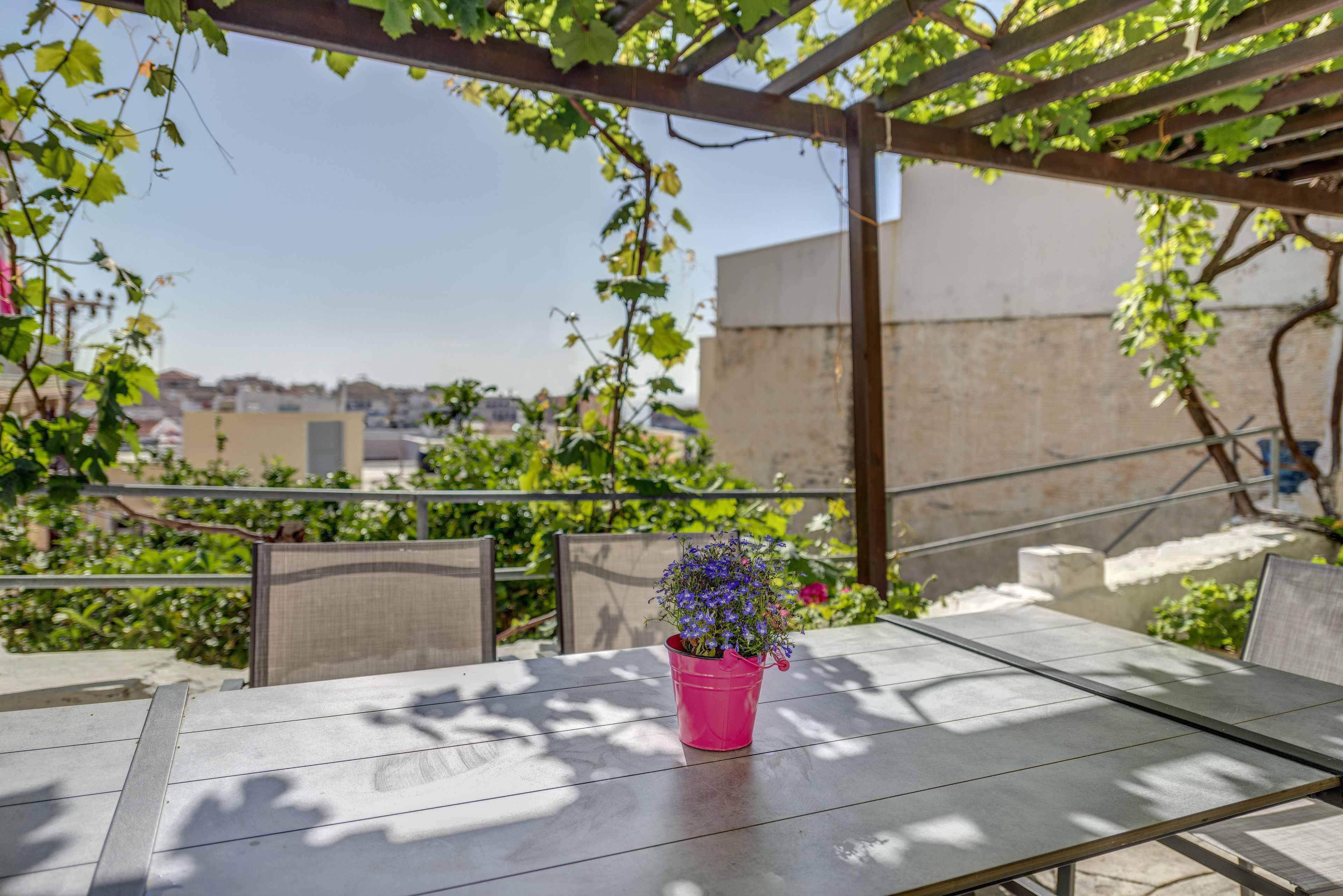 Um pátio com mesa e cadeiras, um vaso de flores cor-de-rosa e vista para edifícios ao fundo.