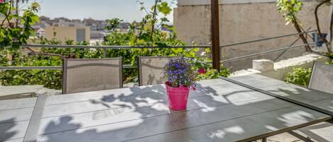Um pátio com mesa e cadeiras, um vaso de flores cor-de-rosa e vista para edifícios ao fundo.