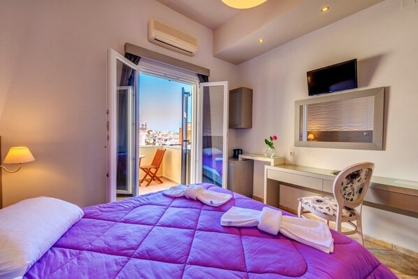 A hotel room with a bed, a desk, a TV, and a balcony with a city view. - Ethrion (Syros)