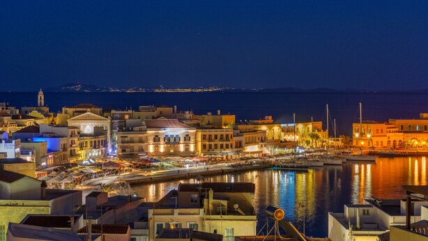 Un paisaje urbano costero nocturno con edificios iluminados, un puerto deportivo y vistas al mar.