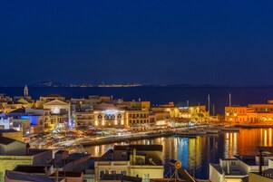 Un paisaje urbano costero nocturno con edificios iluminados, un puerto deportivo y vistas al mar.