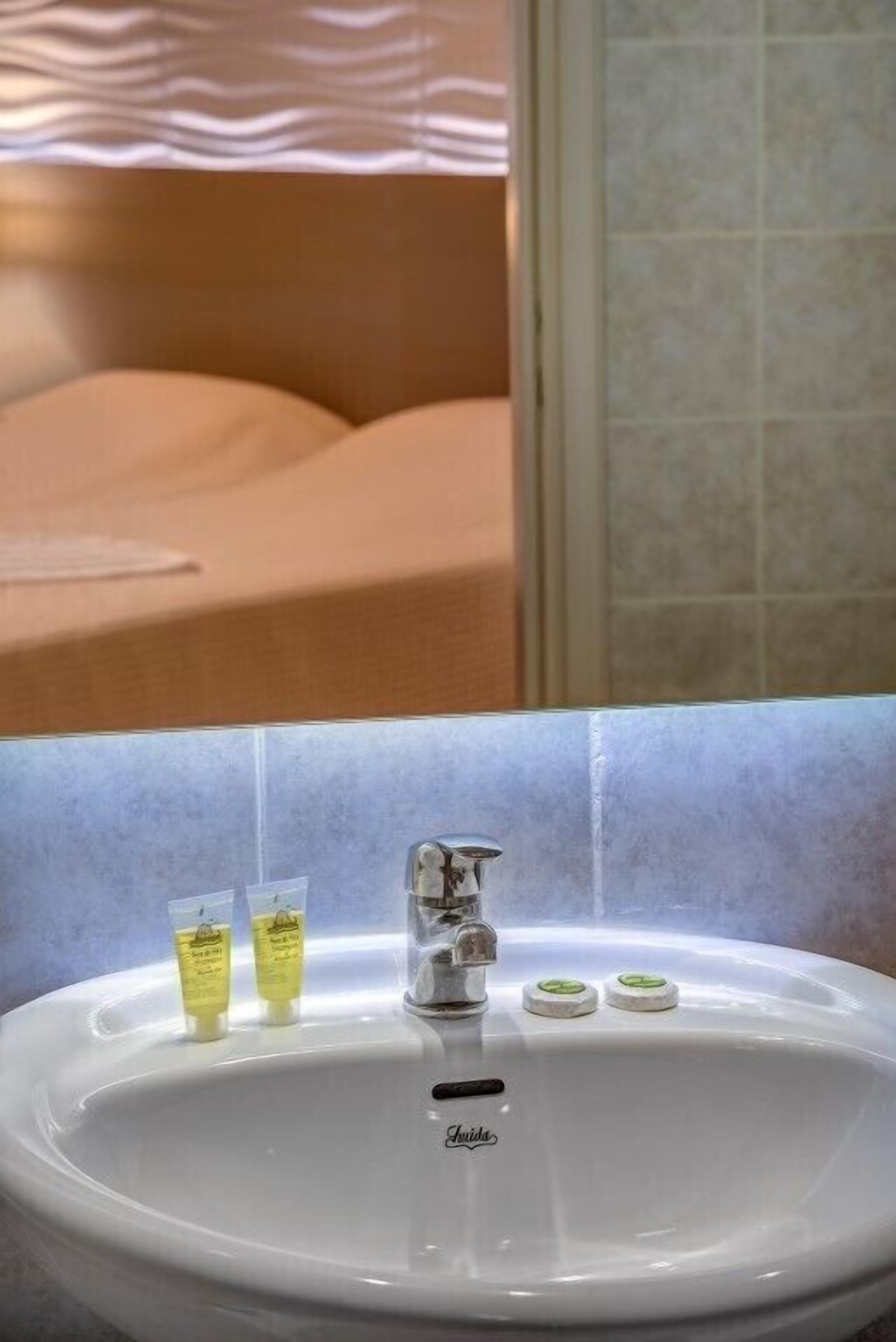 Une salle de bain avec un lavabo blanc, deux petits flacons de lotion et un lit en arrière-plan.