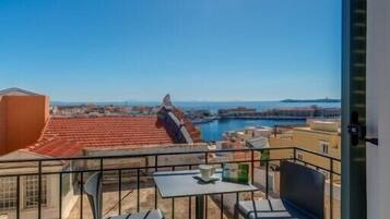 A balcony with a table and chairs, overlooking a coastal area with buildings and a clear sky.