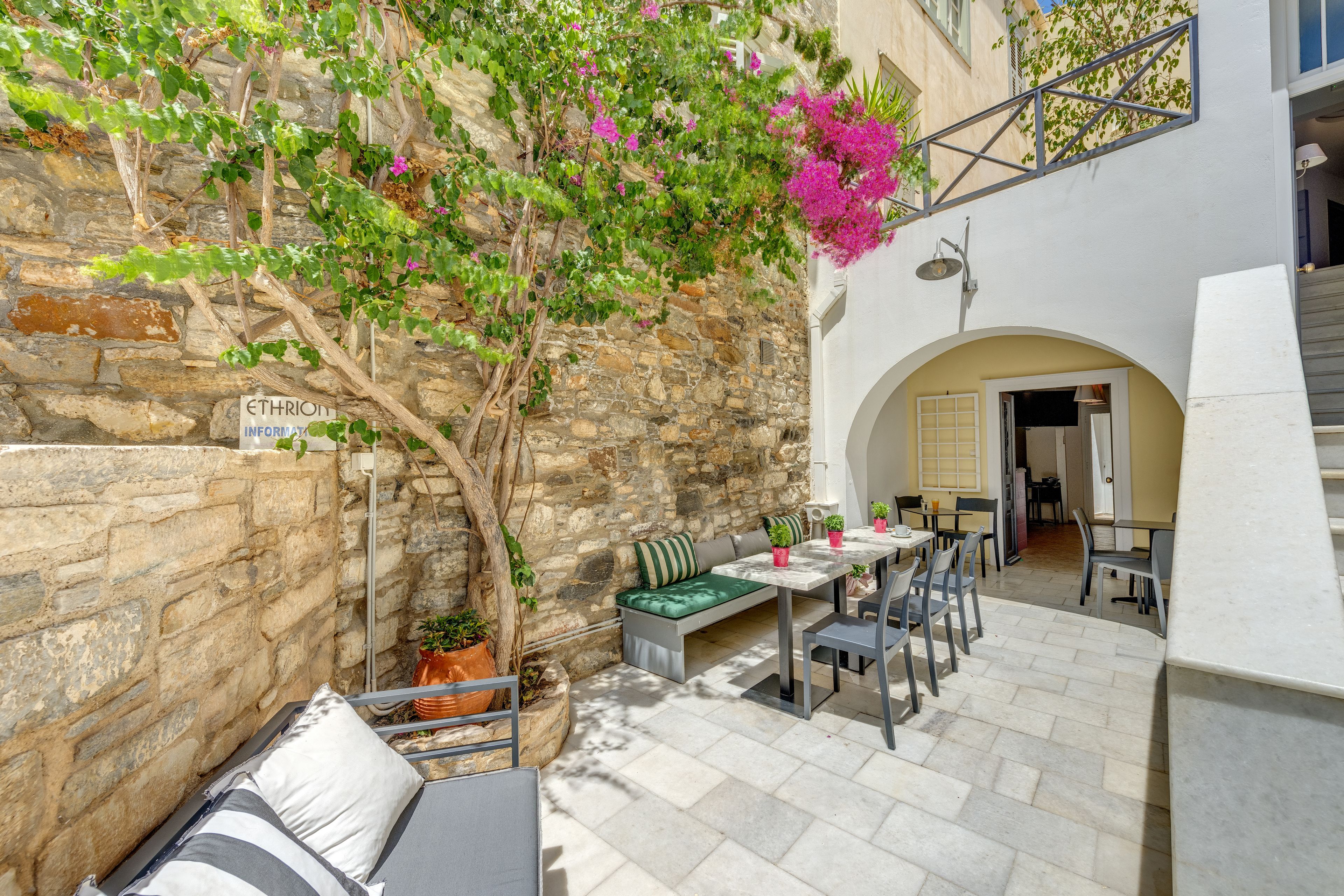A stone-walled outdoor seating area with a bench and tables, surrounded by potted plants and a tree with pink flowers.