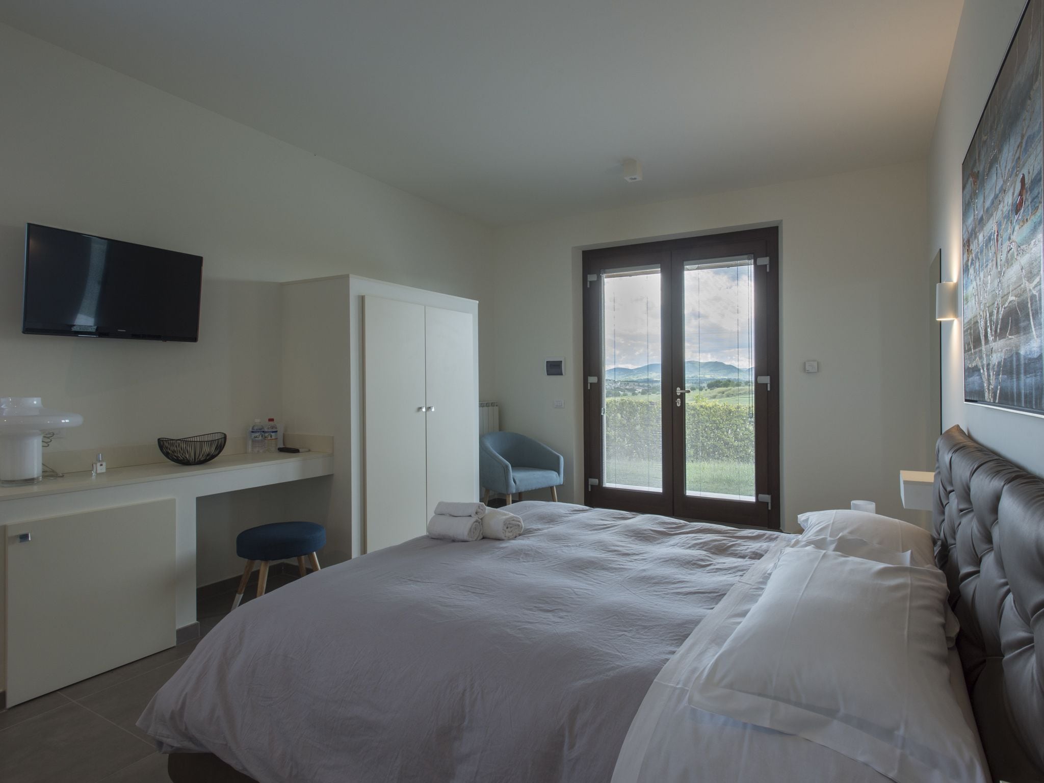 A bedroom with a bed, a desk, a chair, a television, and a door leading to a balcony with a view of the countryside.