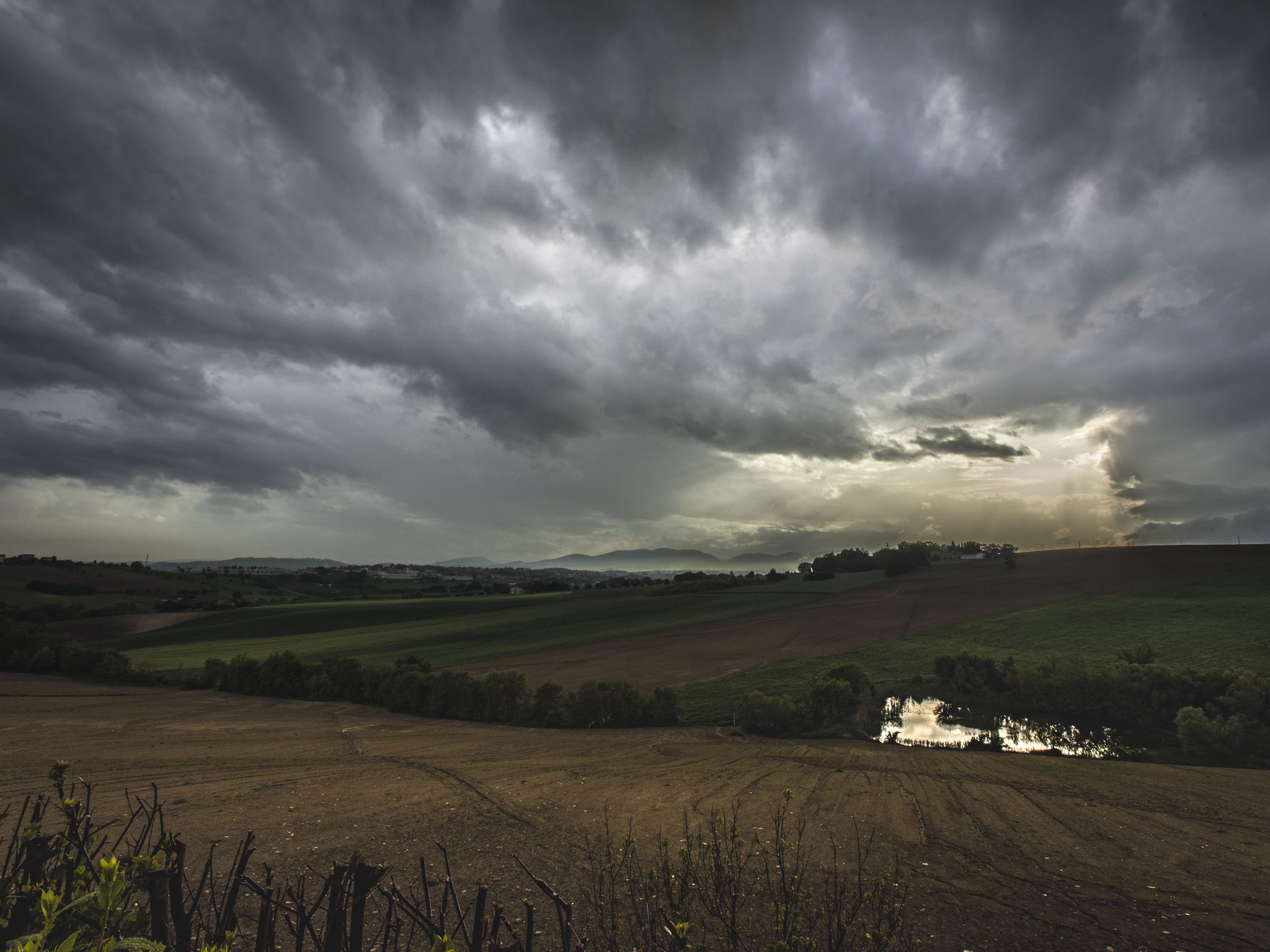 Paisaje rural con campos, un pequeño estanque y cielo nublado.