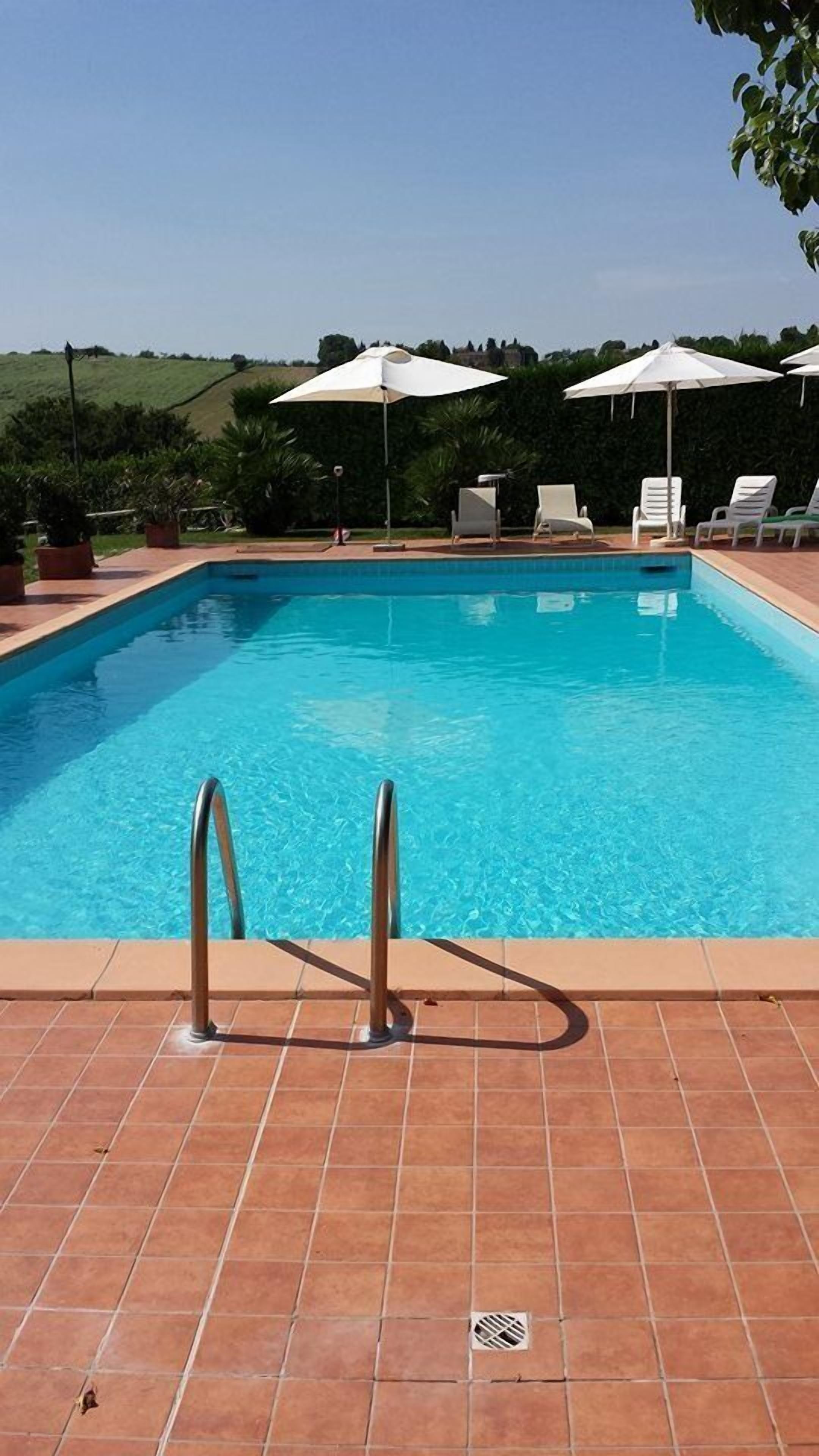 Una piscina rectangular con un borde empedrado, dos escaleras de metal y sombrillas blancas.