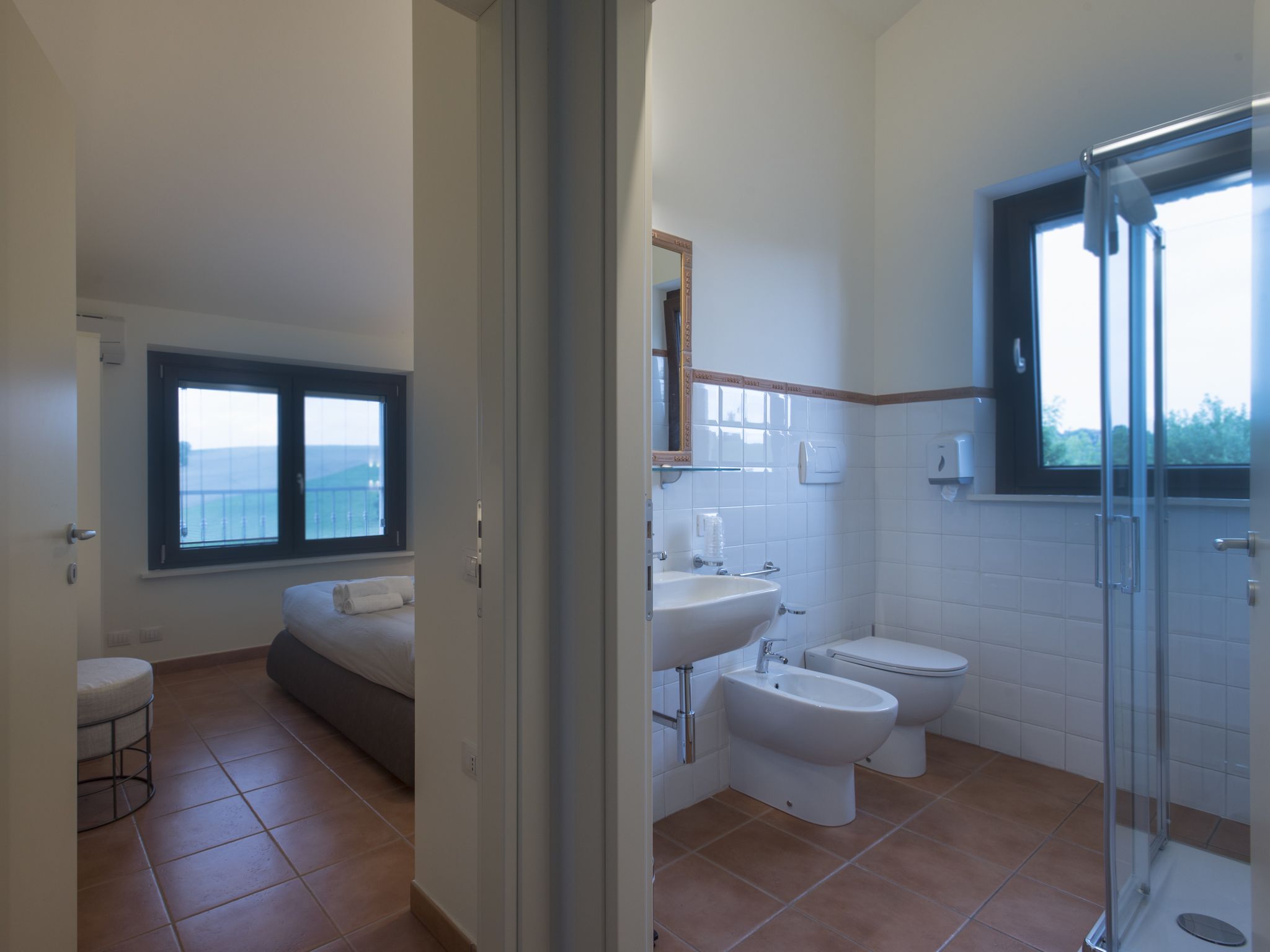Une salle de bain moderne équipée d’une douche, de toilettes, d’un bidet et d’un lavabo. La pièce adjacente contient un lit et une fenêtre.