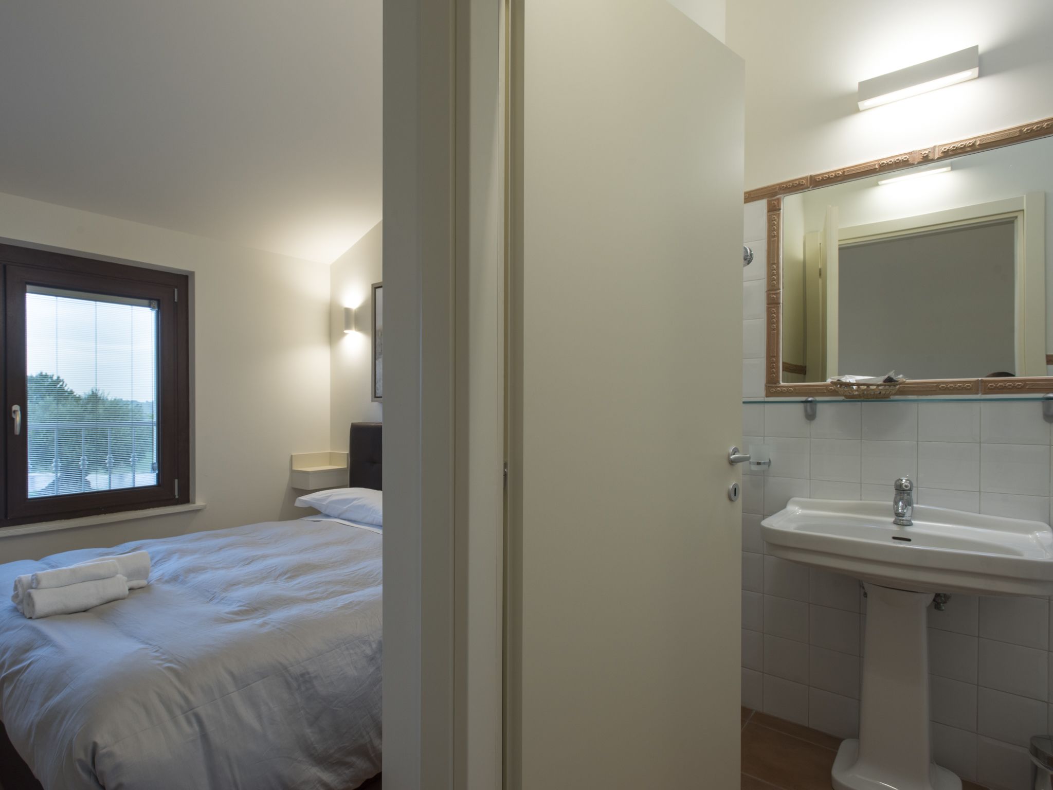 Un dormitorio con cama, ventana, mesita de noche y baño con lavabo y espejo.