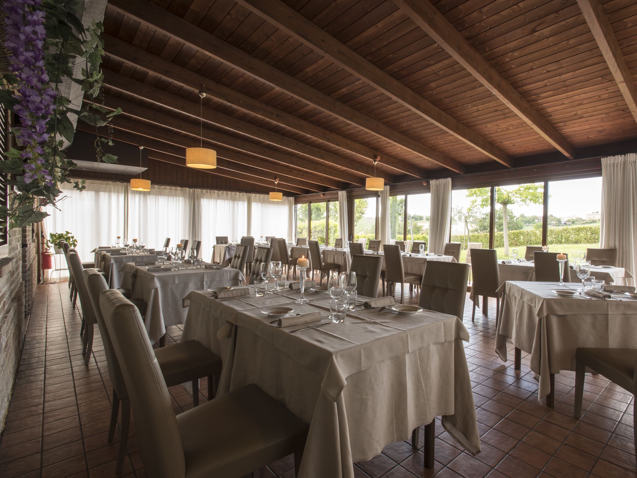 A restaurant with multiple tables set for dining, featuring a wooden ceiling and large windows offering a view of the outdoors.