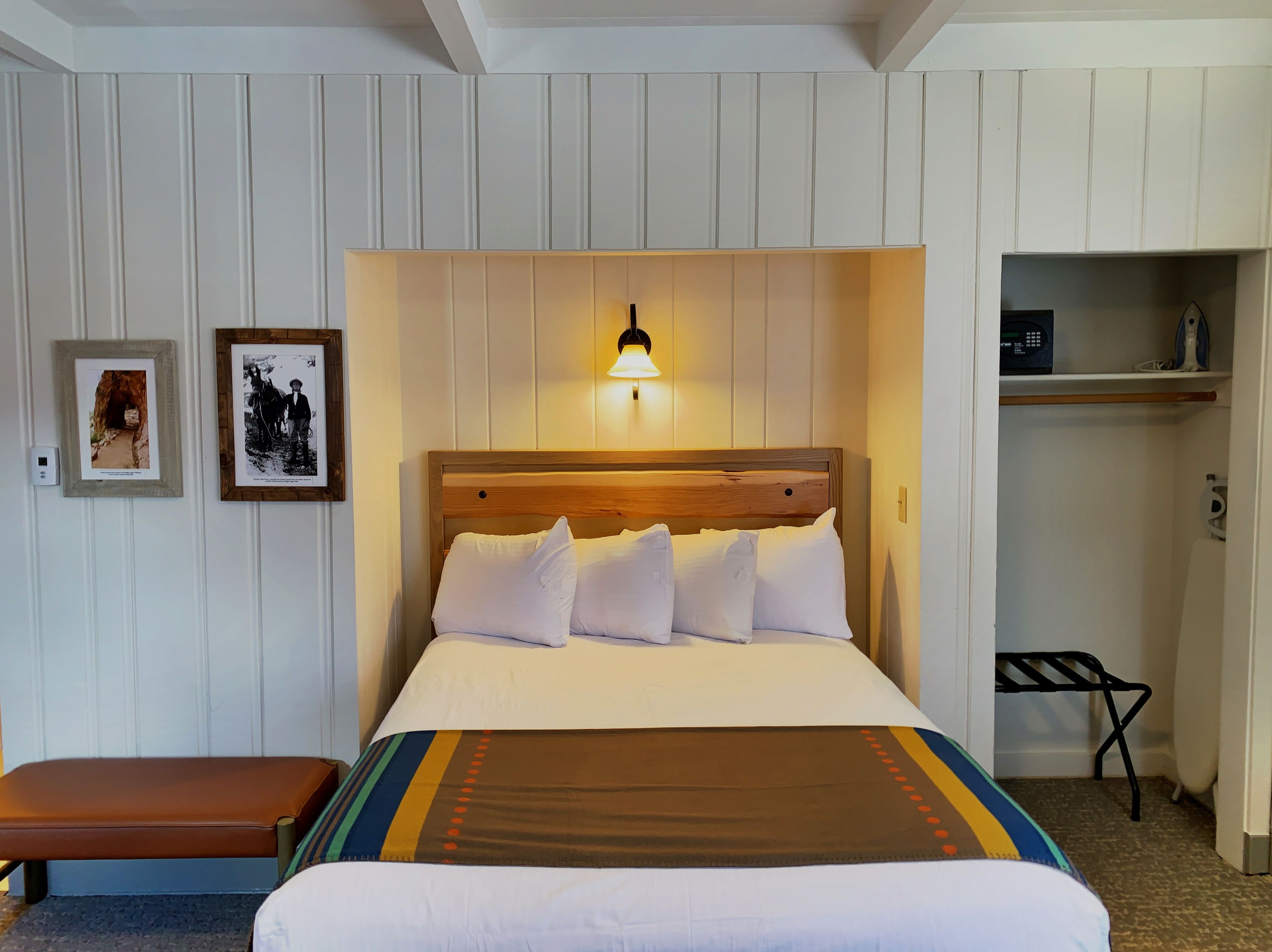 Een netjes opgemaakt bed met witte lakens en een bruine deken, een houten hoofdbord, een wandlamp en een klein plankje met een magnetron en andere spullen.