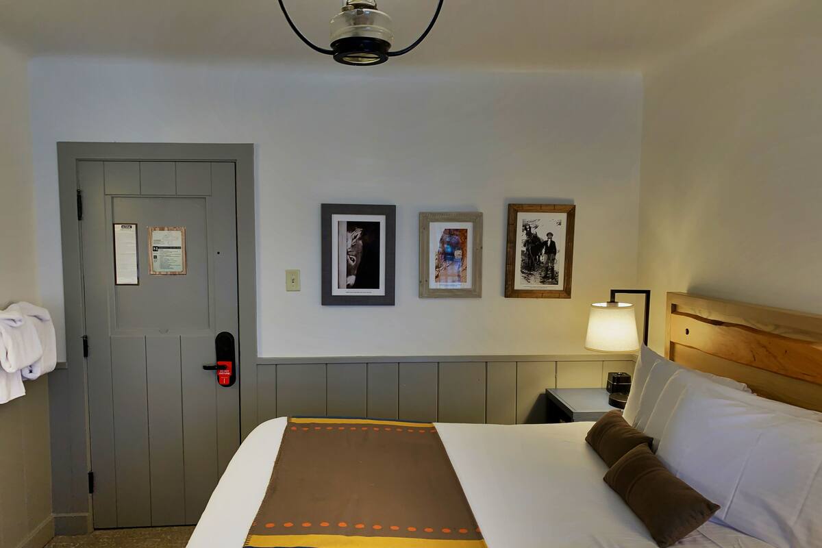 A hotel room with a bed, a door, a wall-mounted lamp, and framed pictures.