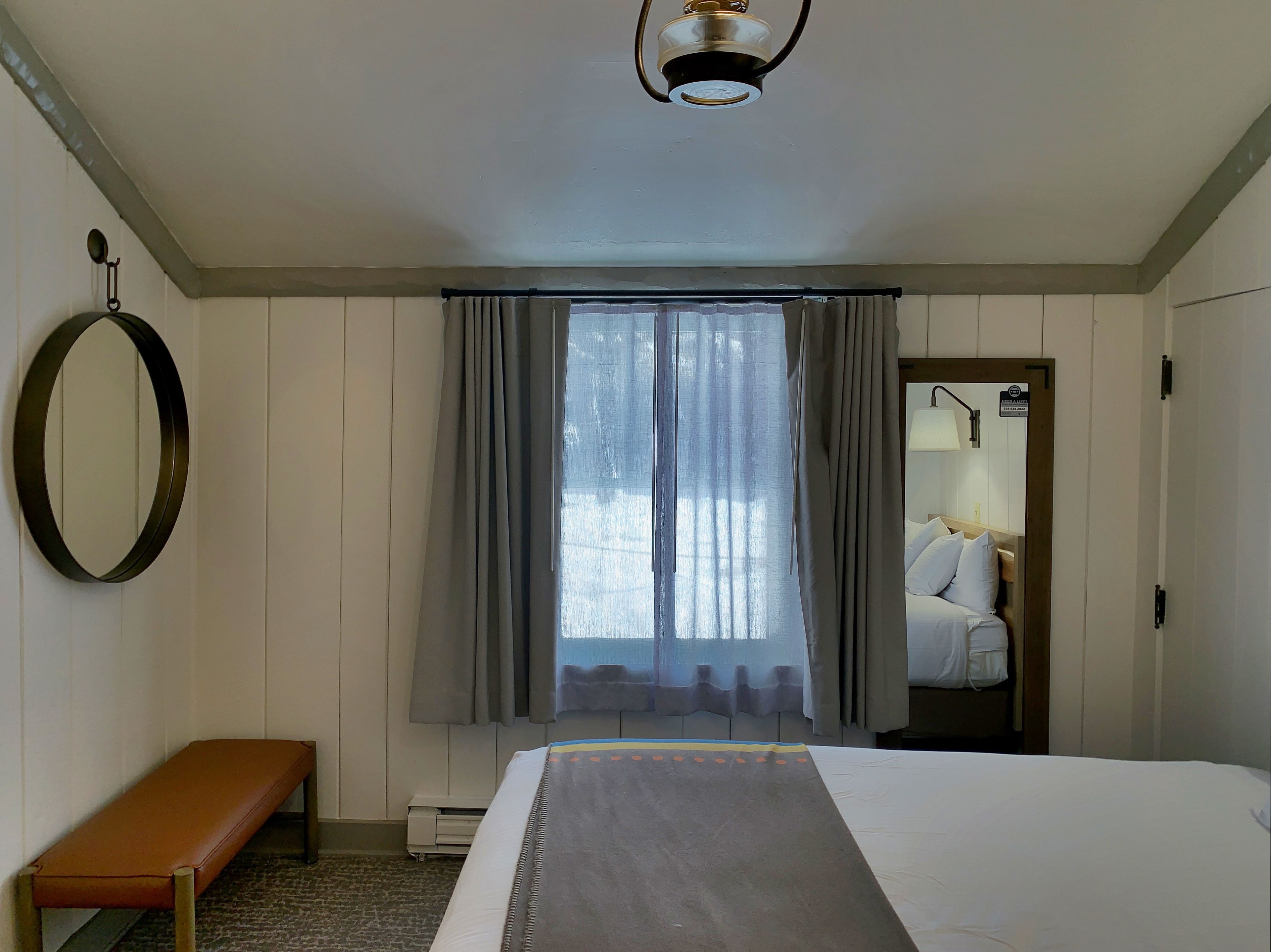 A hotel room with a bed, a bench, a mirror, and a window with curtains.
