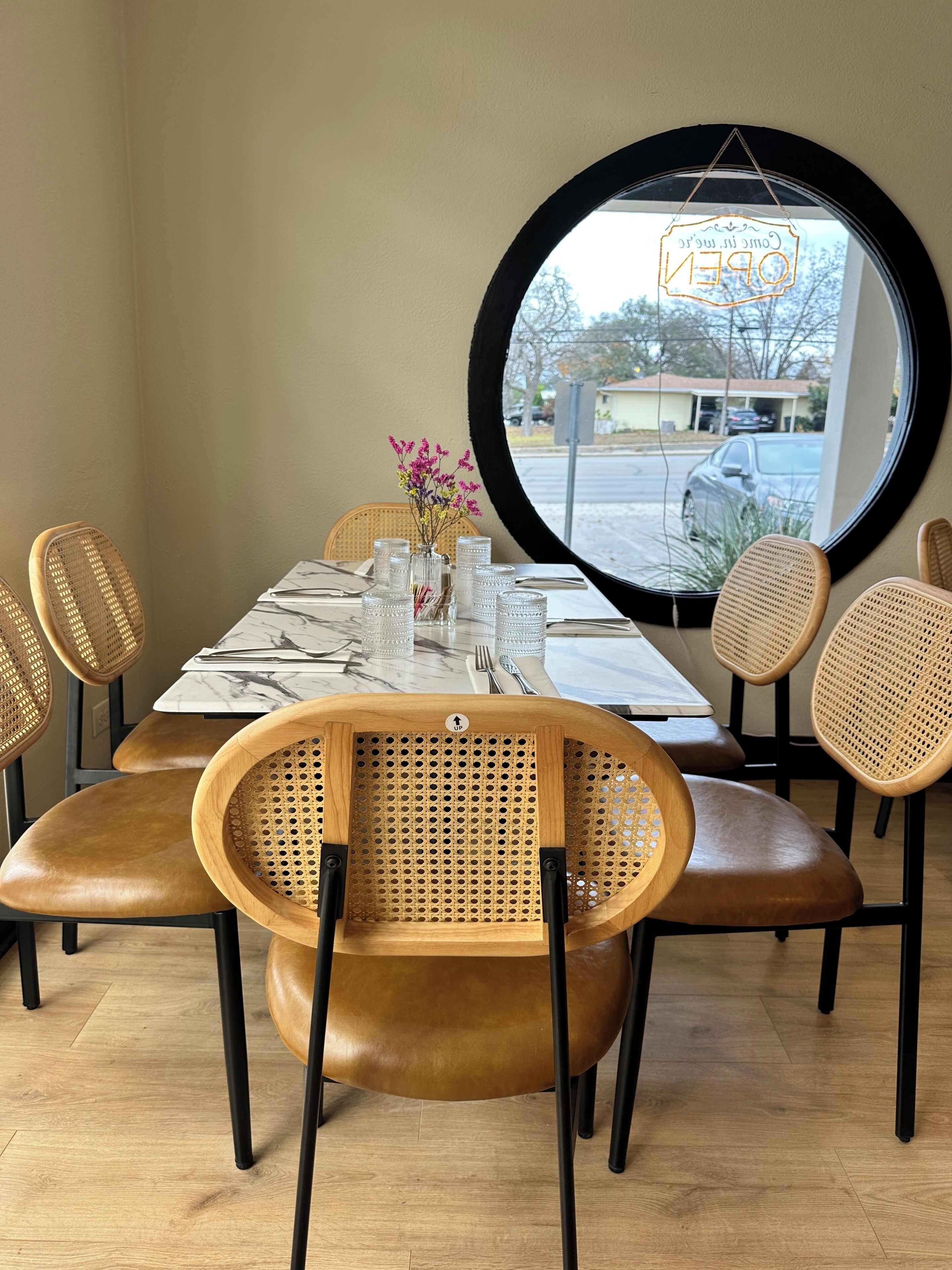 A dining area with a round mirror, wicker chairs, a marble table, and a view of the outside.