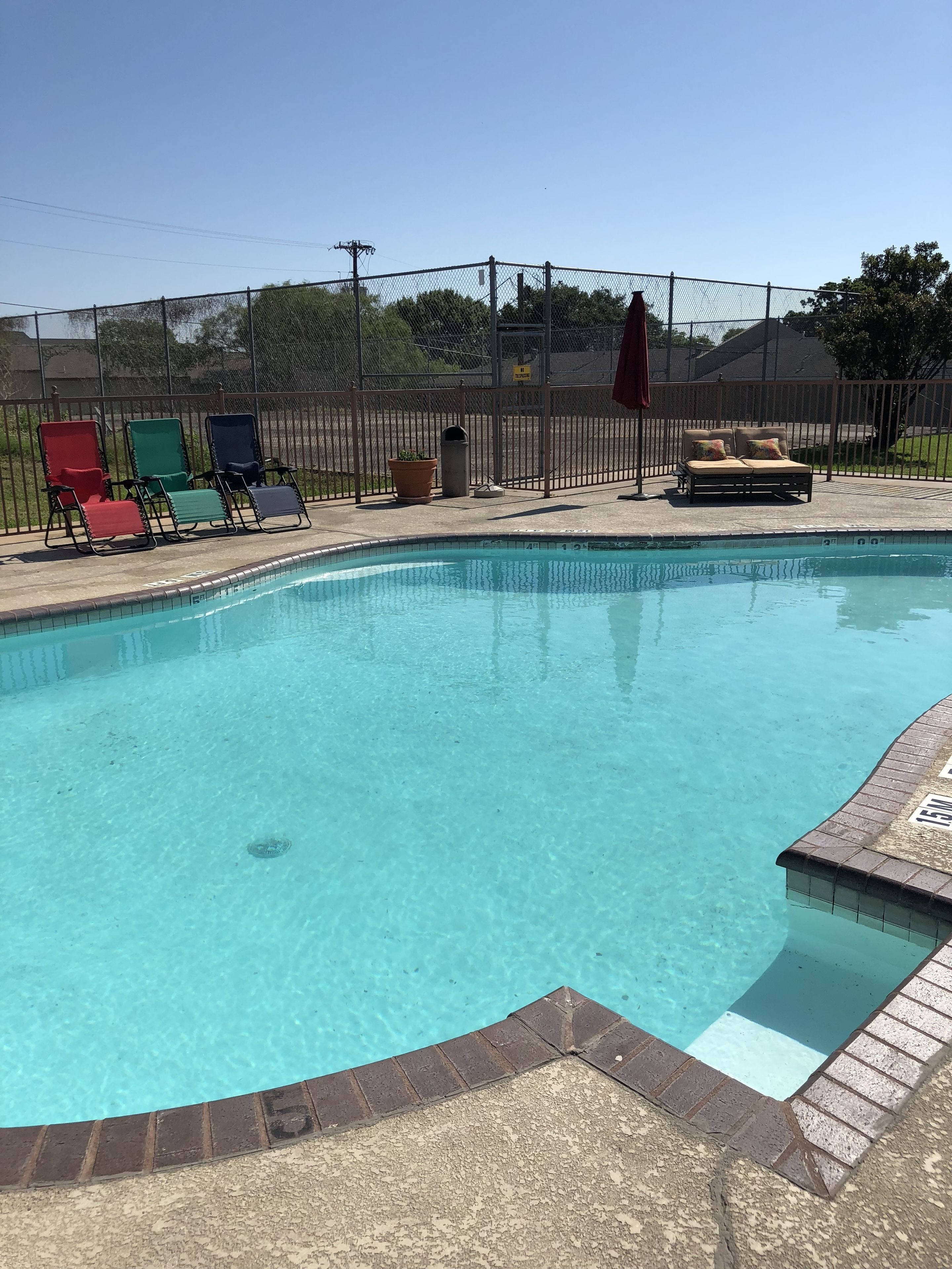 A clear swimming pool with a brick border, surrounded by a fence and various lounge chairs.