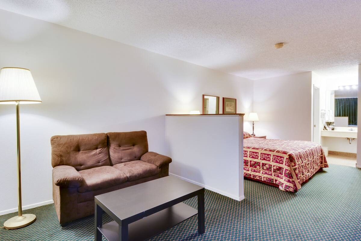 Une chambre d’hôtel avec un canapé marron, une petite table, un lit recouvert d’une courtepointe à motifs, une lampe sur pied et une table de chevet avec une lampe.