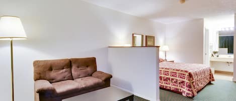 Habitación de hotel con un sofá marrón, una mesita pequeña, una cama con colcha estampada, una lámpara de pie y una mesita de noche con lámpara.