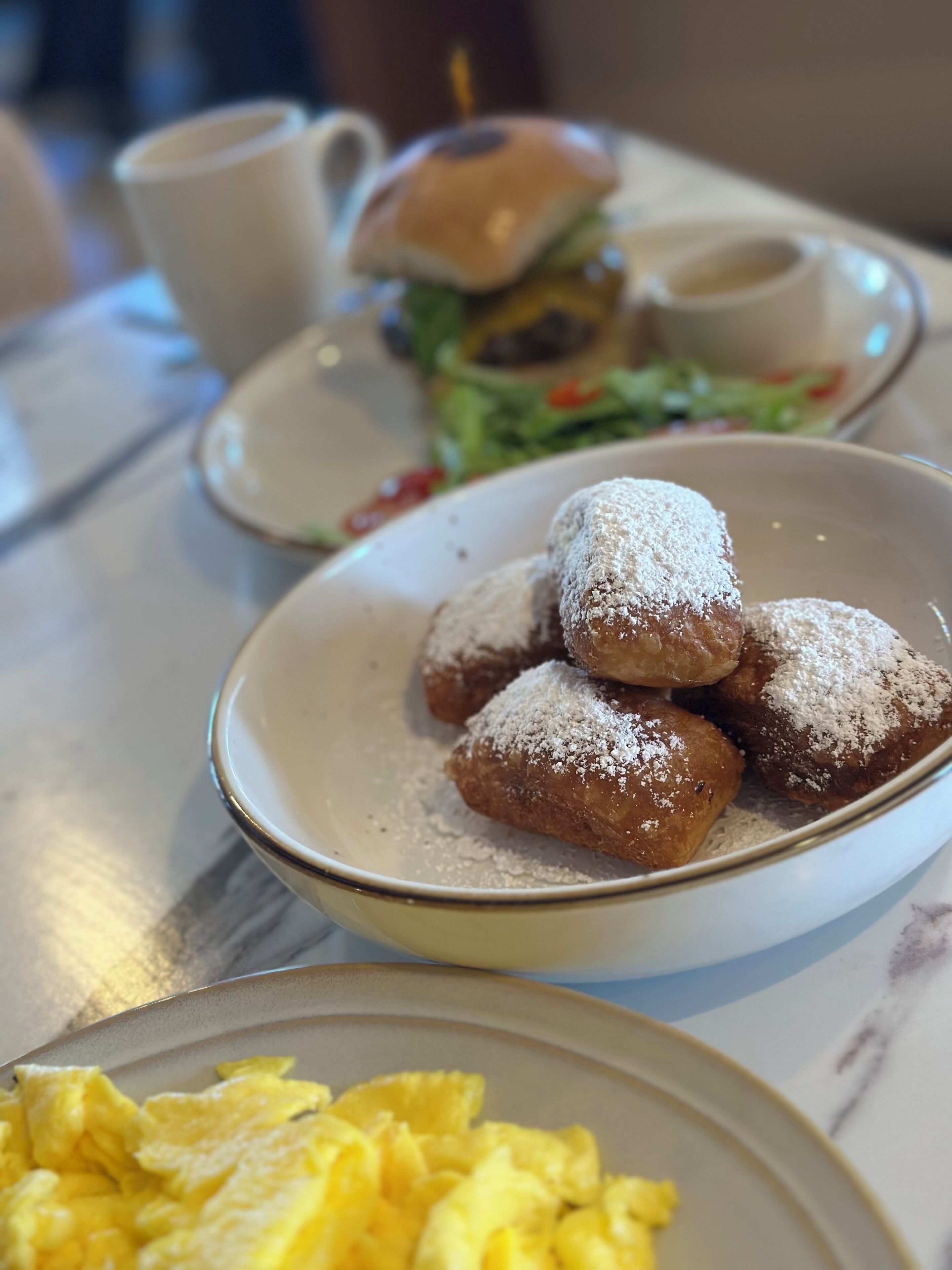 A plate of powdered doughnuts, a burger with a bun, and scrambled eggs on a table.