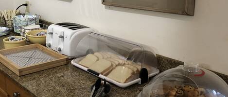 Una encimera de cocina con un tostador, pan, un panero, un cortador de pan y un recipiente con pasteles.