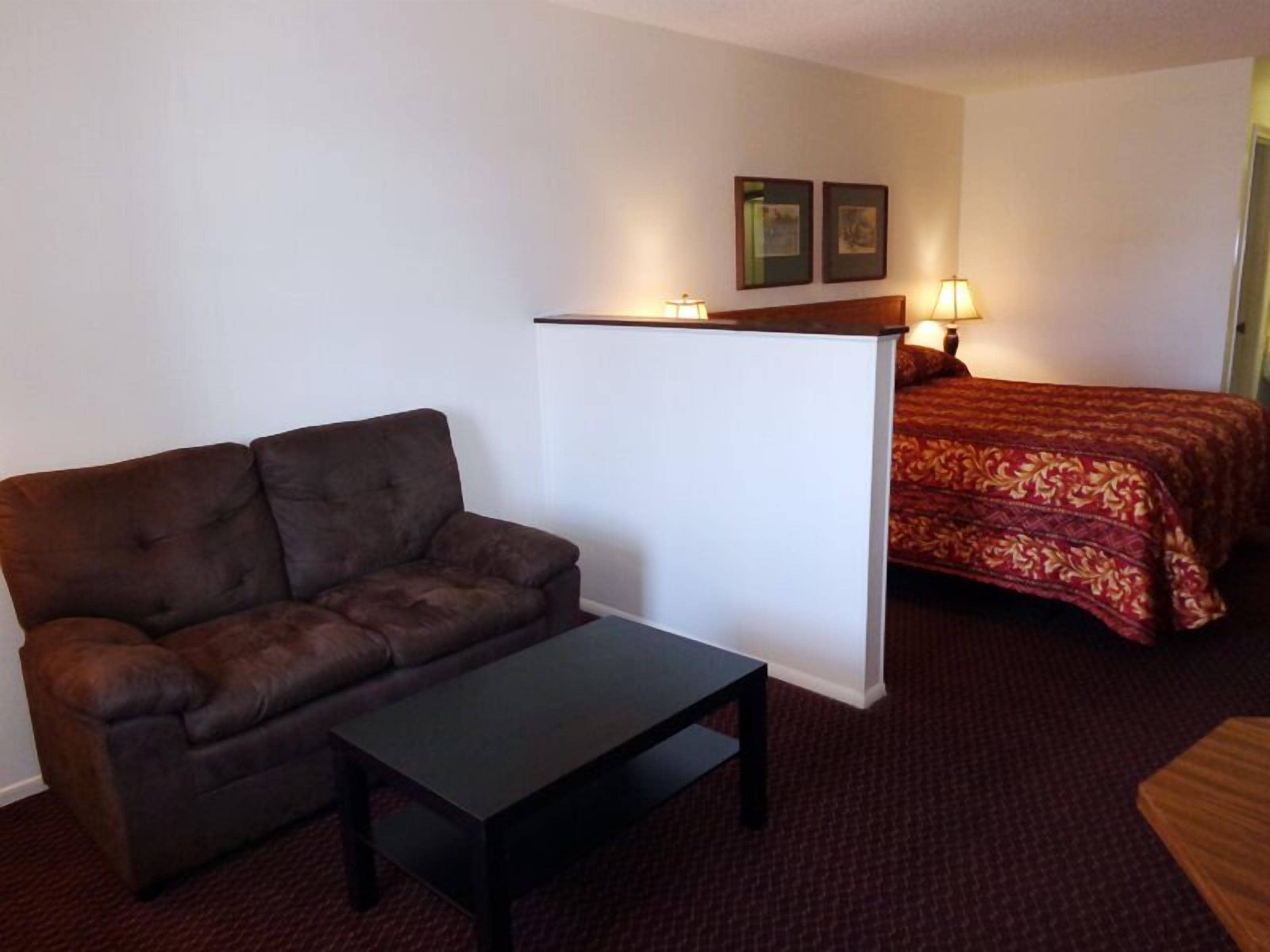 A hotel room with a brown sofa, a small table, a bed with a red patterned bedspread, and two wall-mounted lamps.