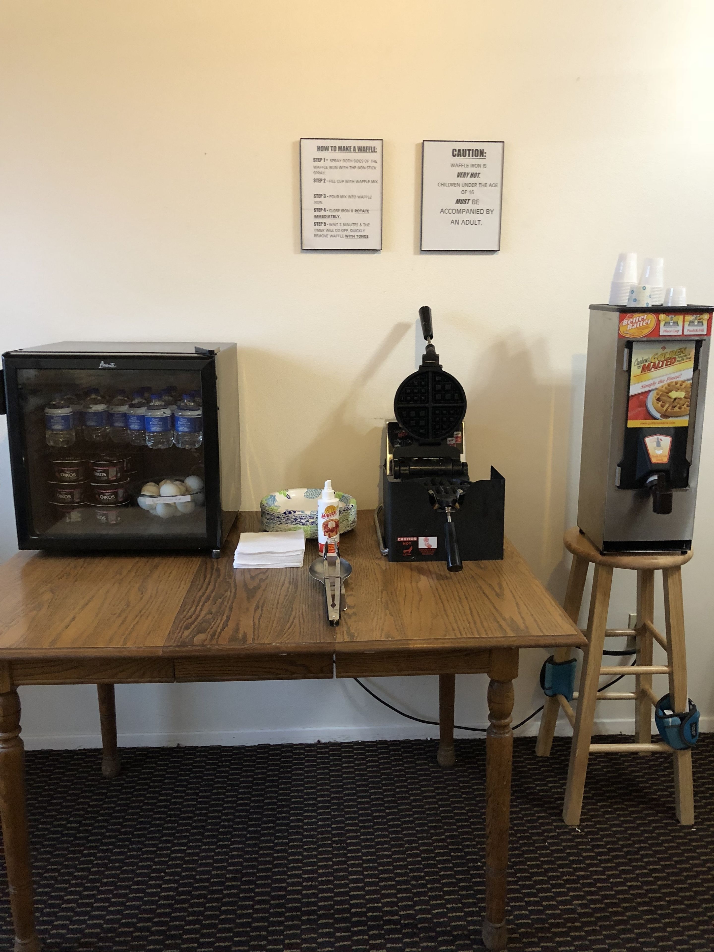 A table with a waffle iron, a bottle of syrup, and a waffle maker. There are also two signs on the wall and a refrigerator with beverages.