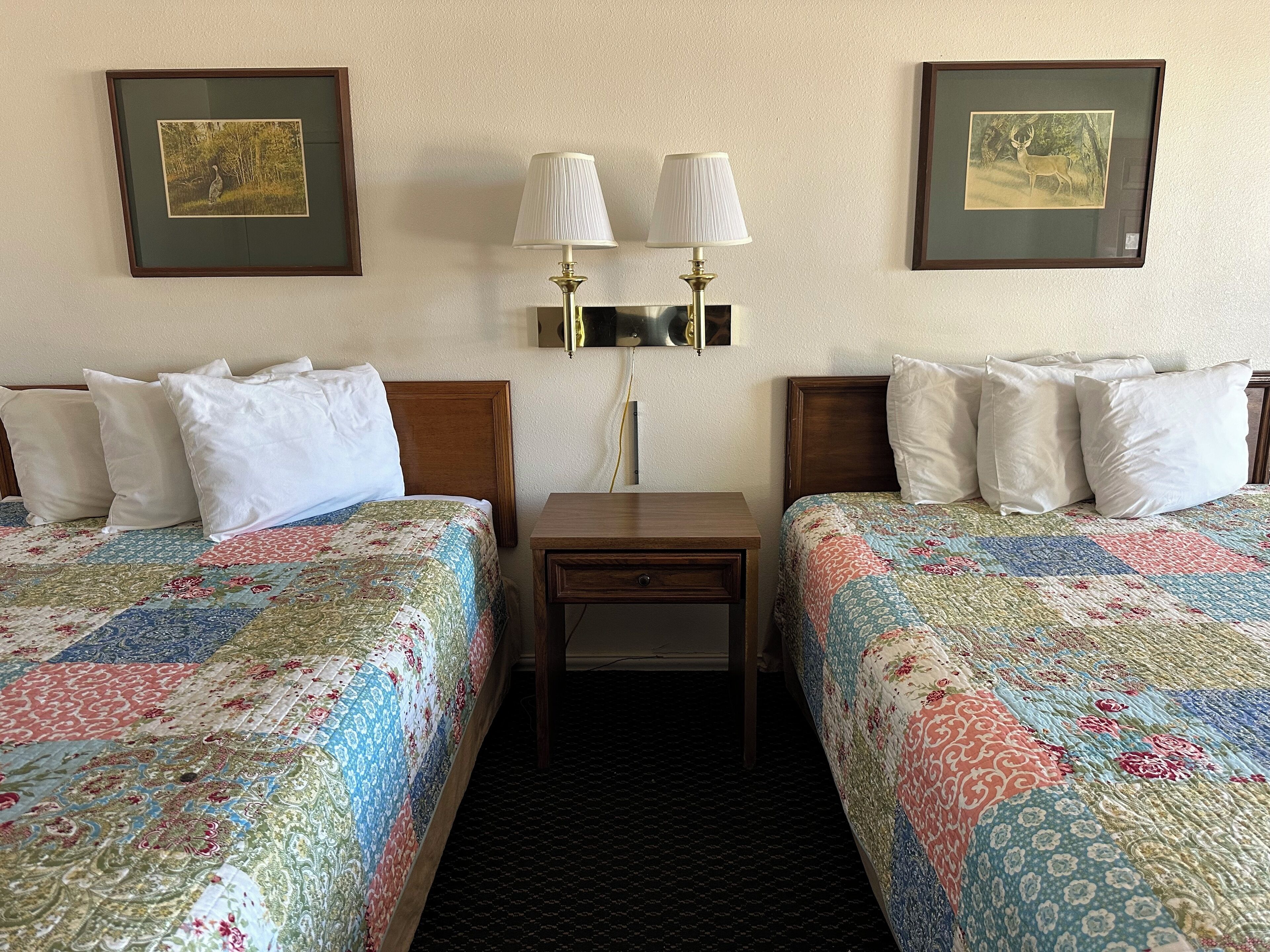 A hotel room with two beds, each with a floral quilt, a nightstand, a lamp, and framed pictures on the wall.