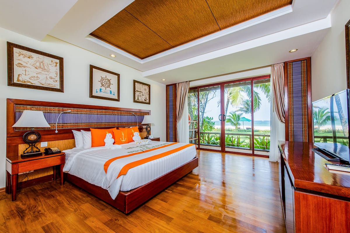 Une chambre spacieuse avec un grand lit, une tête de lit en bois et une vue sur la plage grâce à de grandes fenêtres.