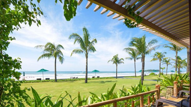 Um deck de madeira com guarda-corpo, com vista para uma praia, palmeiras e céu claro.
