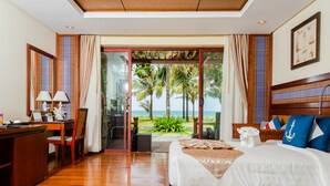 Quarto de hotel com chão de madeira, escrivaninha com luminária, cama com almofadas e vista para palmeiras e o mar.