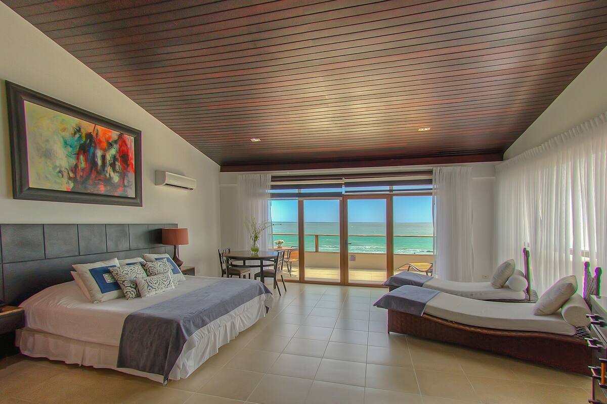 A hotel room with a bed, a sofa, a dining table, and a view of the ocean.