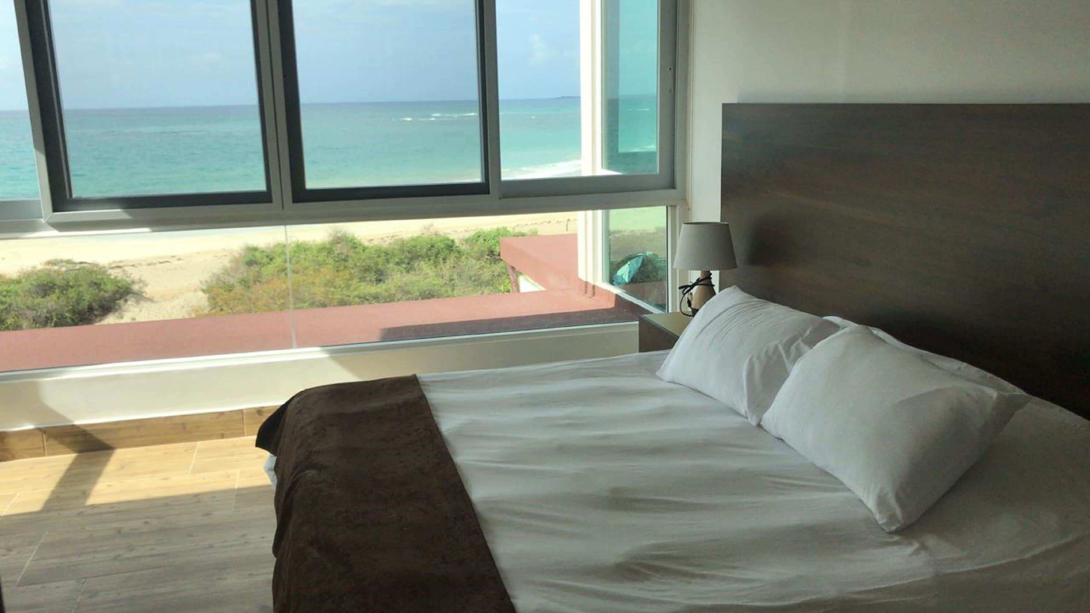 A bedroom with a large window overlooking a beach and ocean.