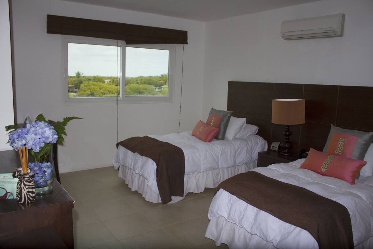 A hotel room with two beds, a window with a view, a bedside table with a lamp, and a decorative vase with flowers.