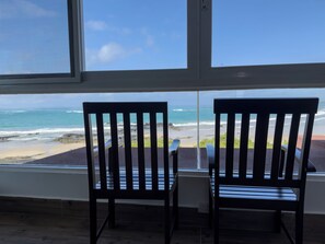 Two wooden chairs facing a window overlooking a beach with waves and a clear sky.