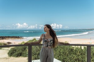 Una persona parada en un balcón con vistas a una playa, cielo despejado y olas del océano.