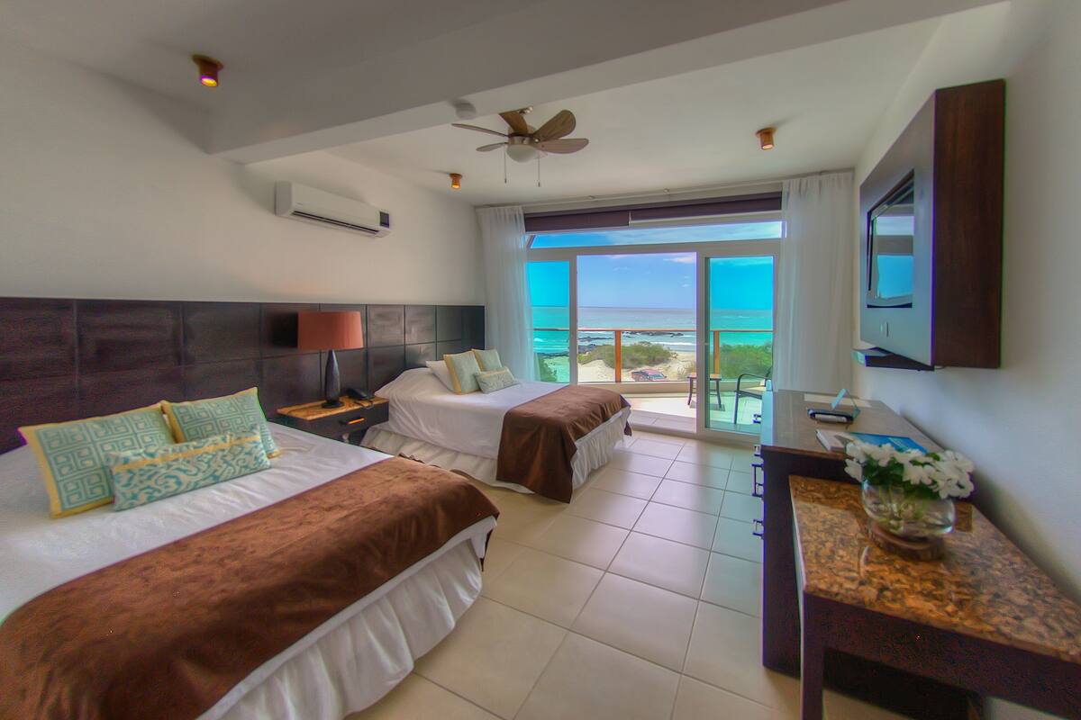A hotel room with two beds, a desk, a ceiling fan, and a large window with a view of the ocean.