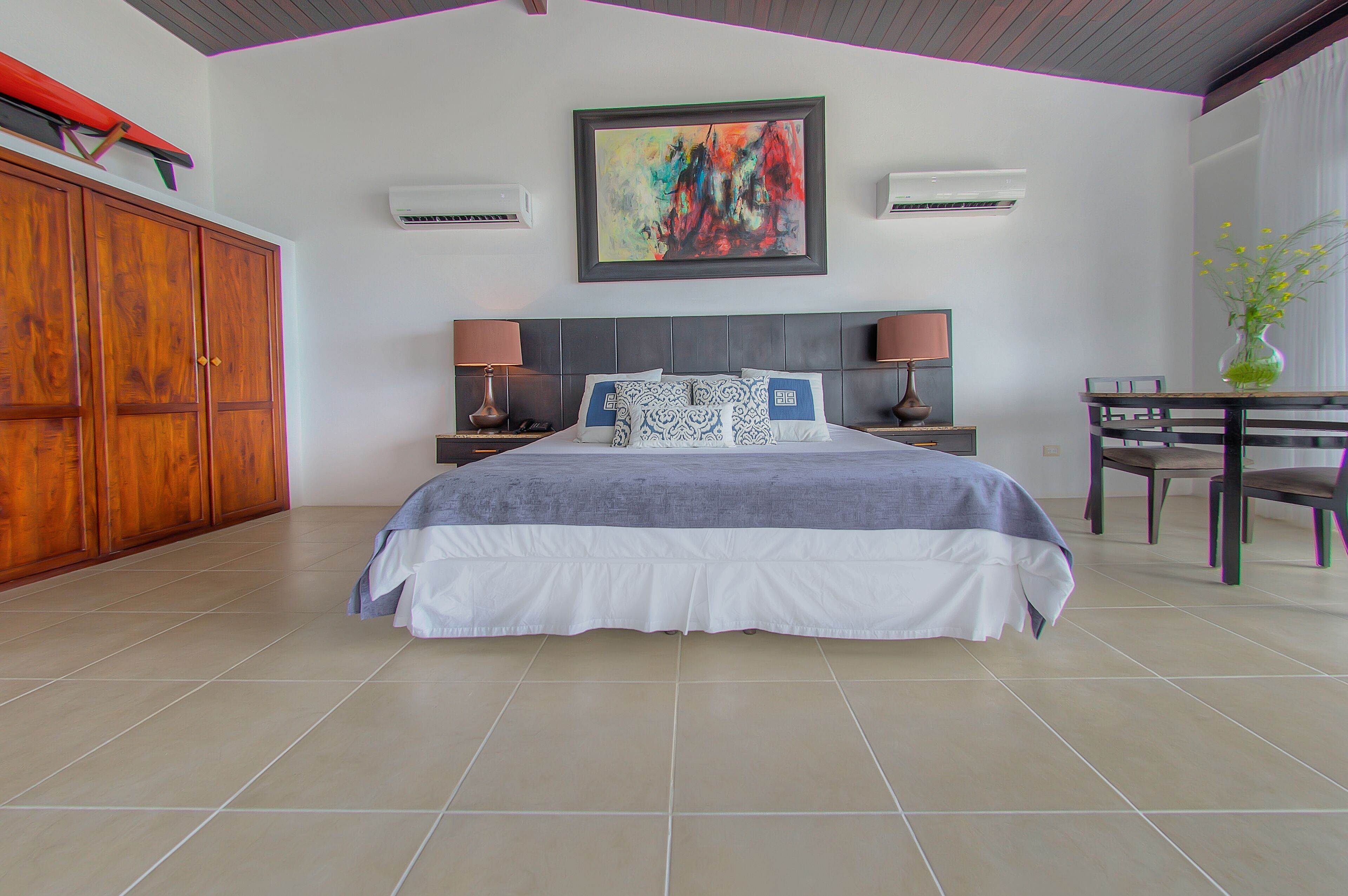 A bedroom with a large bed, two bedside tables with lamps, a wooden wardrobe, a dining table with chairs, and a colorful abstract painting on the wall.