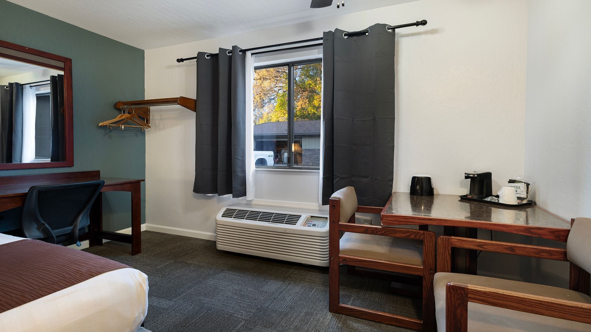 A hotel room with a bed, a desk, a chair, a heater, a window with curtains, and a view of the outside.