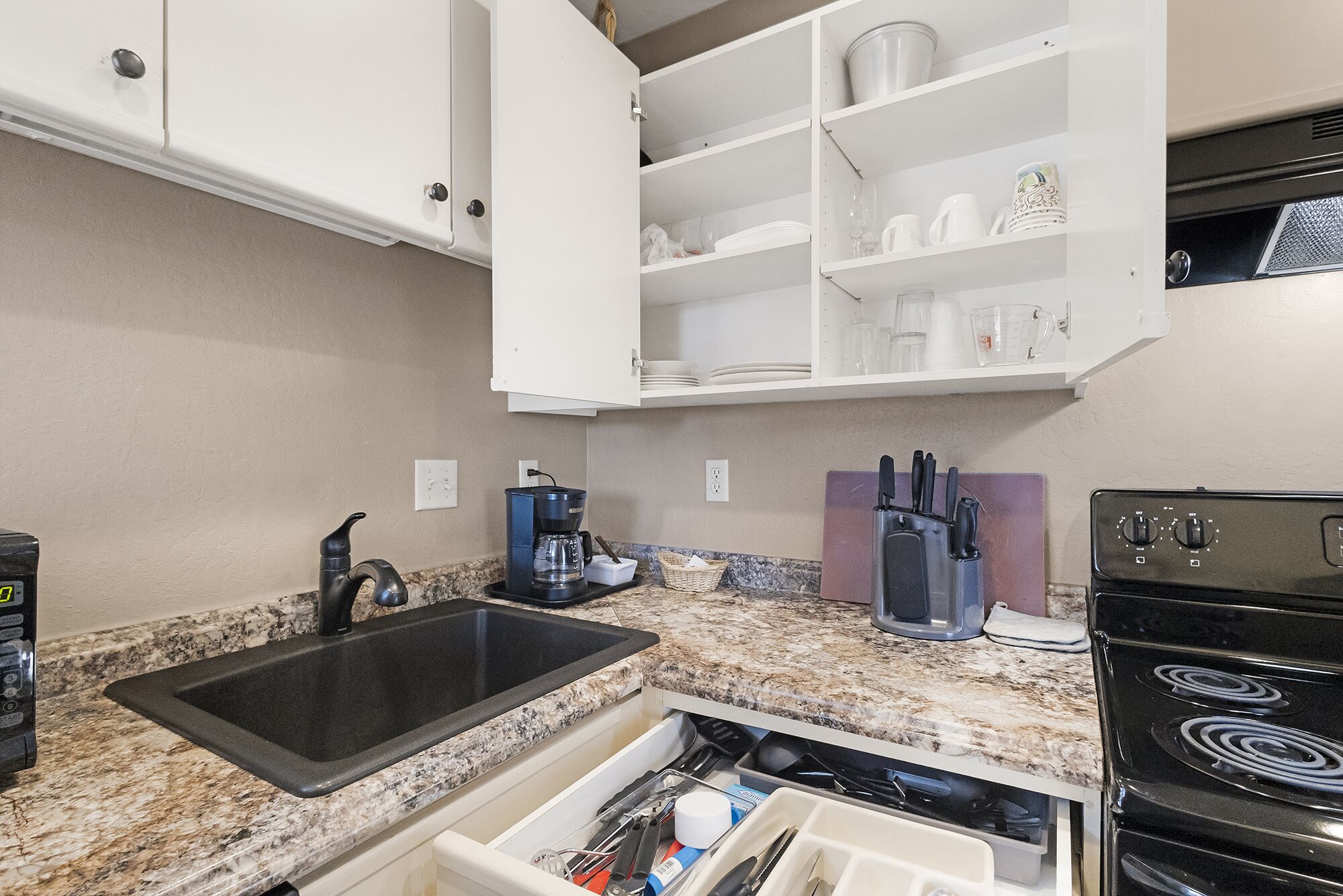 A kitchen with a granite countertop, a black sink, a coffee maker, a stove, and white cabinets.