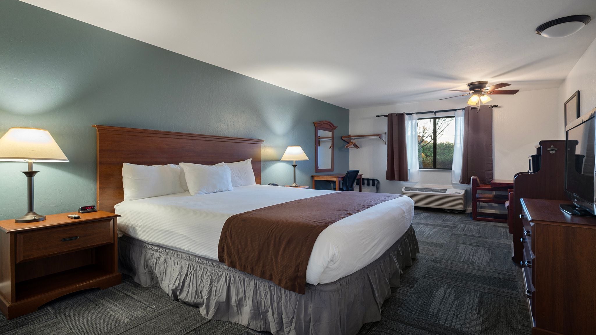 A hotel room with a large bed, two bedside tables with lamps, a desk with a chair, a TV, and a ceiling fan.