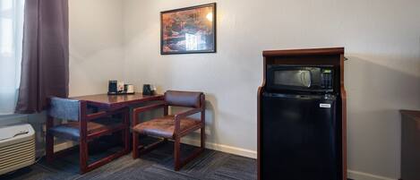 A compact hotel room with a kitchenette, a dining table with two chairs, a refrigerator with a microwave, and a painting on the wall.
